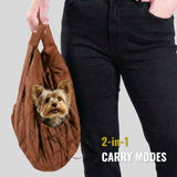 Small dog in brown 2-in-1 pet carrier sling bag, person holding handles, urban dog gear