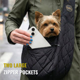 Small dog in a quilted black pet carrier sling with two large zipper pockets, woman holding phone.
