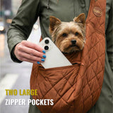 Small dog in brown quilted pet carrier sling with large zipper pocket holding a phone, urban setting