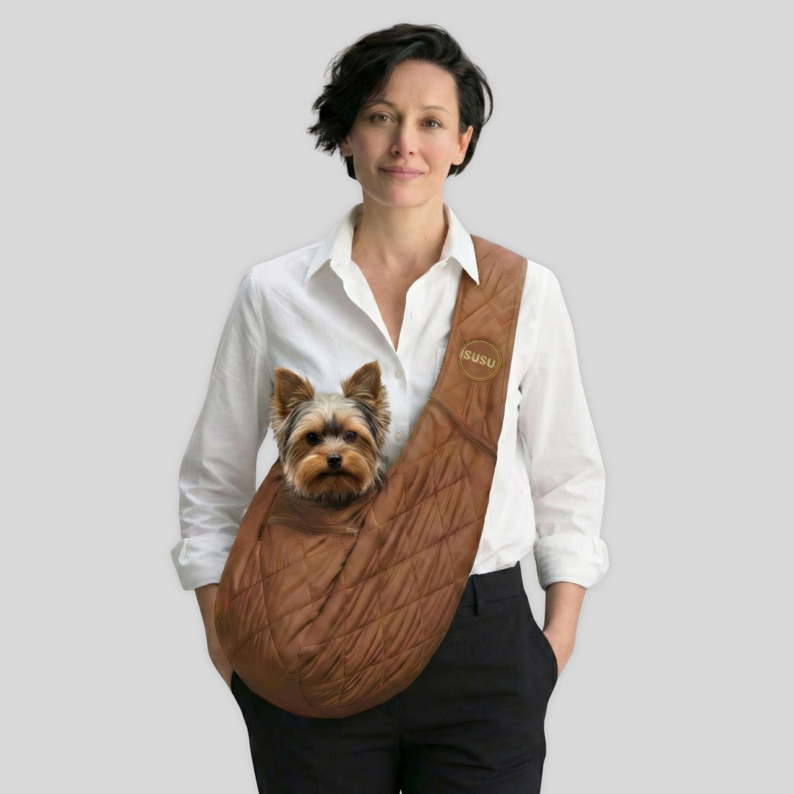 Woman carrying a small dog in a brown quilted pet carrier sling, standing against light background.