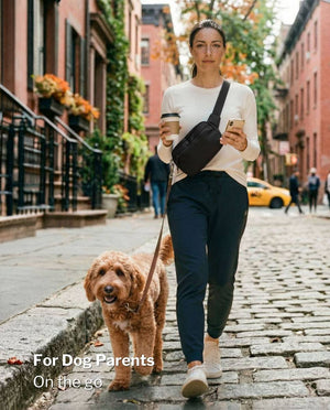 Woman holding coffee and phone while walking her dog, black SUSU Grab & Go dog walking bag worn crossbody on a city street