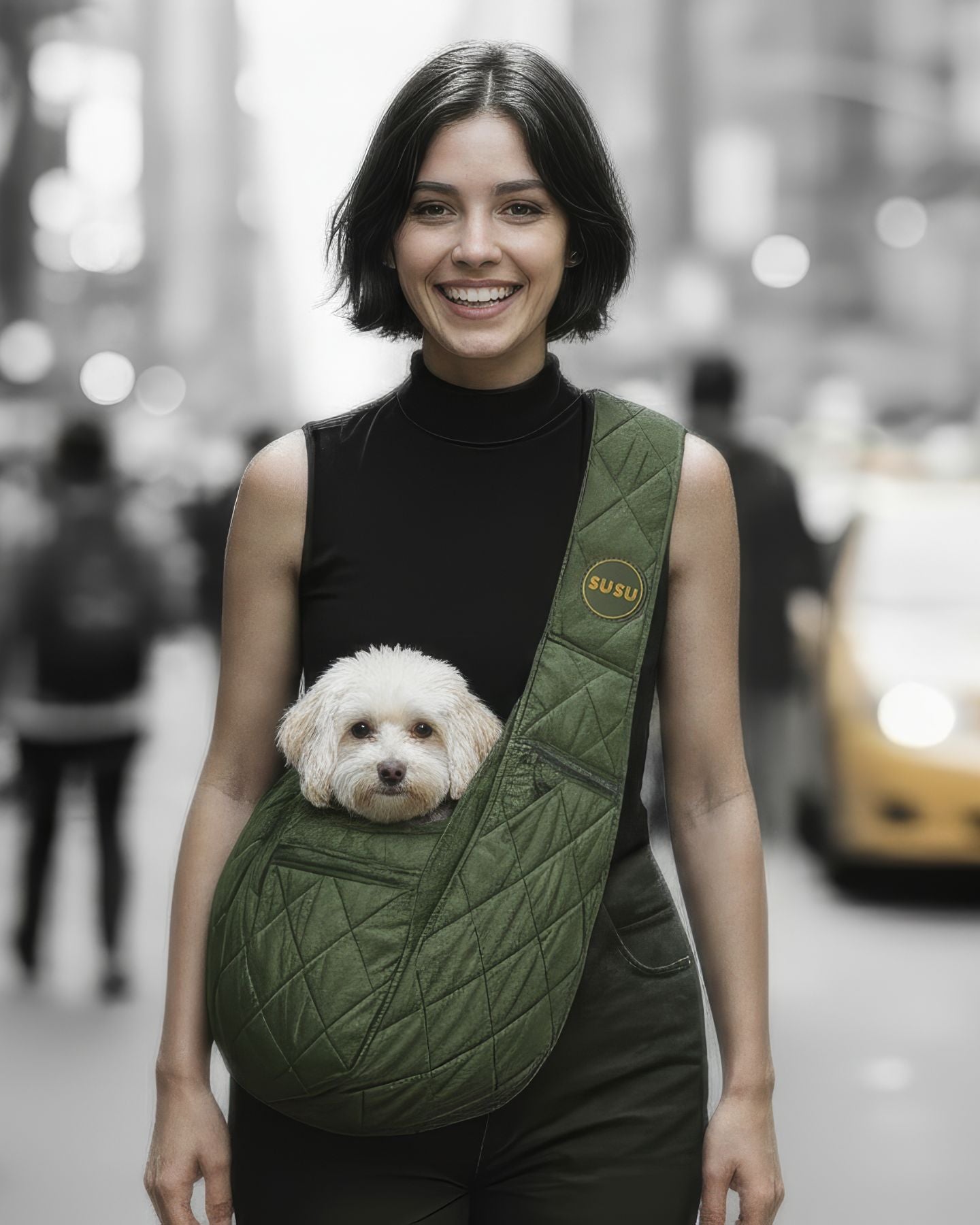 Woman walking in NYC wearing an olive green SUSU small dog carrier sling with a white pup snug in front, blurred city street and taxis behind