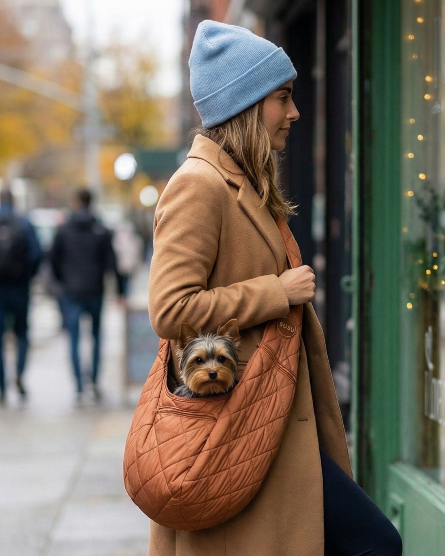 Woman in a camel coat wearing a camel SUSU small dog sling carrier, with a Yorkie peeking out near a storefront